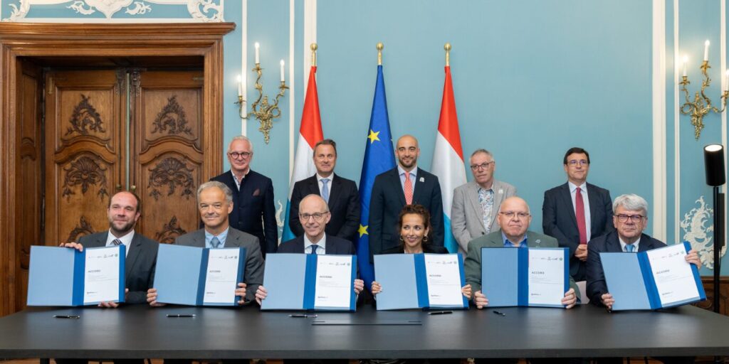 Huit personnes sont assises à une table, des documents ouverts sous les mains, tandis que cinq autres se tiennent debout derrière elles dans une salle solennelle ornée de drapeaux et de portes en bois sculptées.