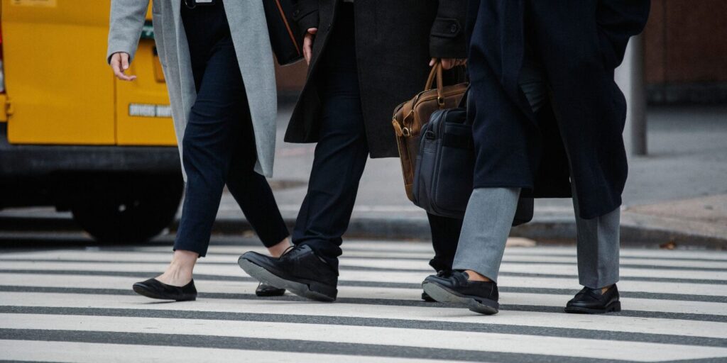 Trois personnes, vêtues de manteaux et portant des sacs, traversent un passage piéton en milieu urbain.